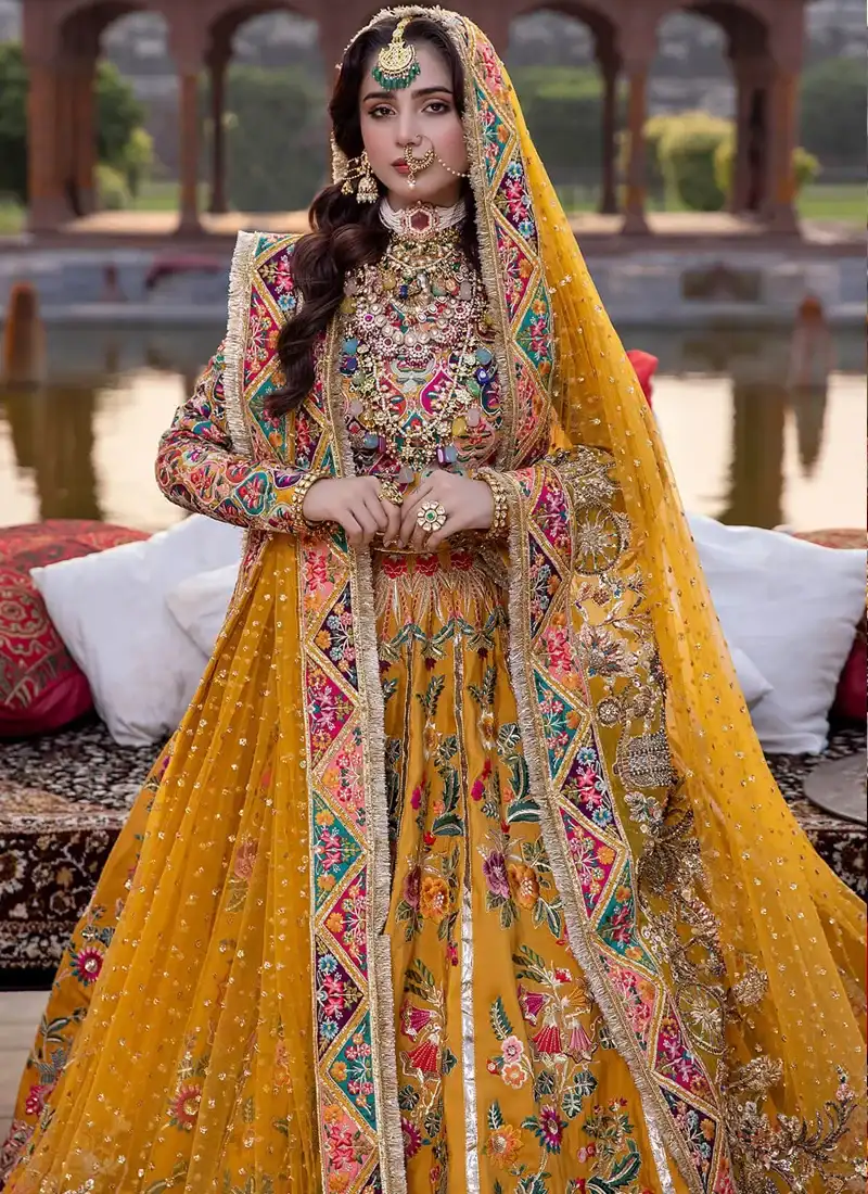 Mustard lehenga choli seated angled view showing pink and teal sleeve motifs and floral skirt panels